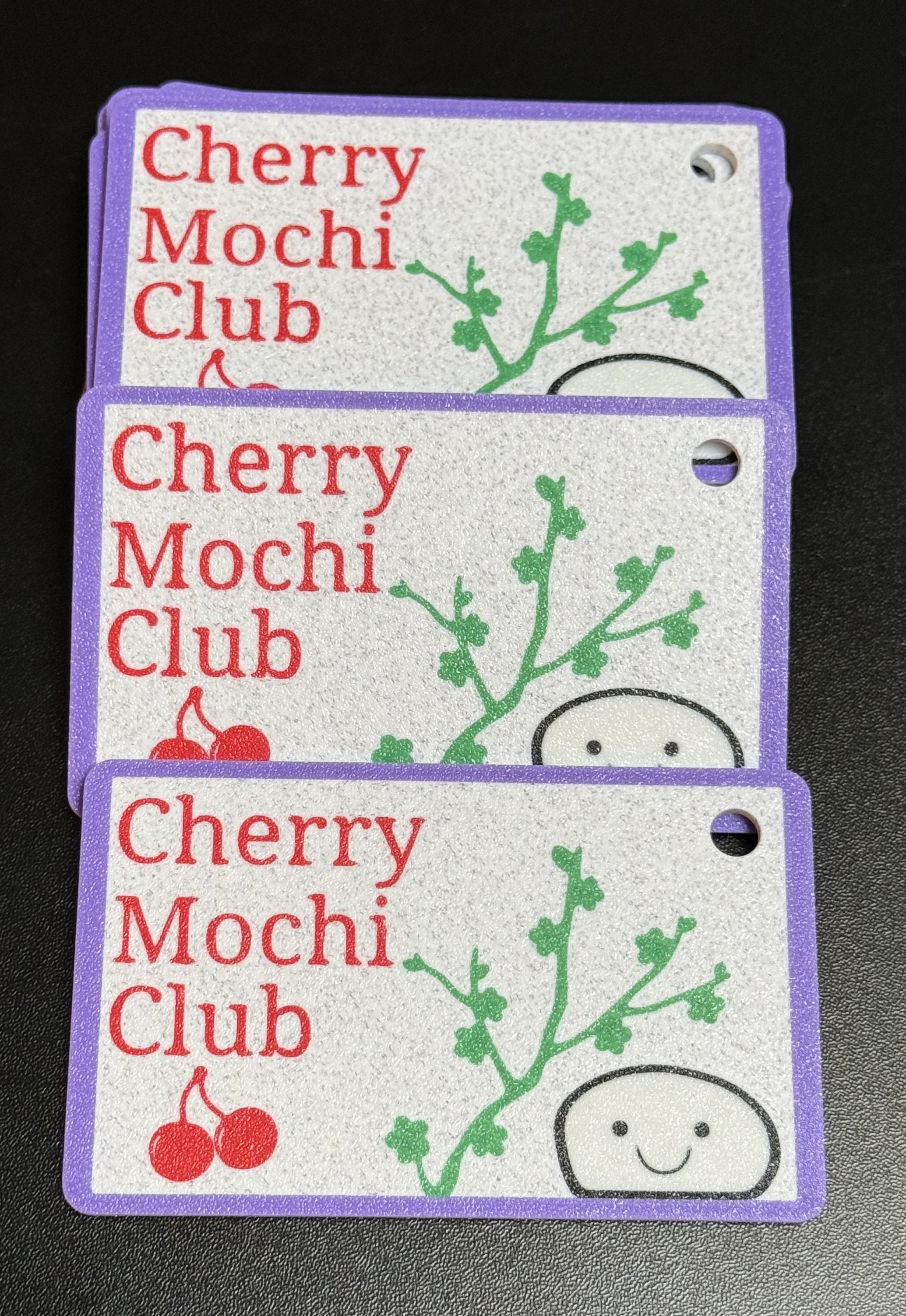 A small stack of 3d printed cards, with the text Cherry Mochi Club on each of them, along with a styalized cherry, cherry blossom branch, and a piece of mochi with a smiling face on it.