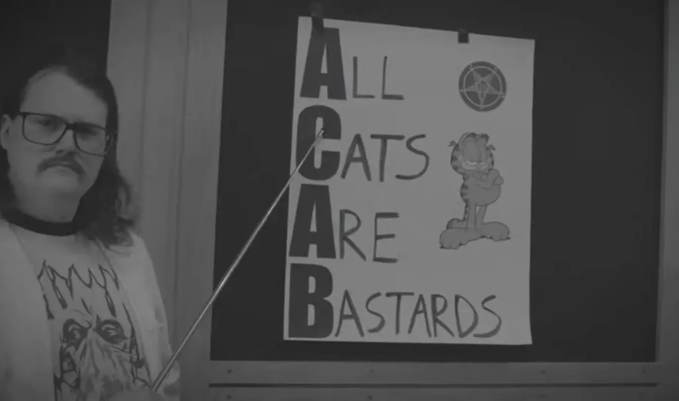 A man holding a stick pointing to a poster with the words 'All Cats Are Bastards' written next to a picture of Garfield.