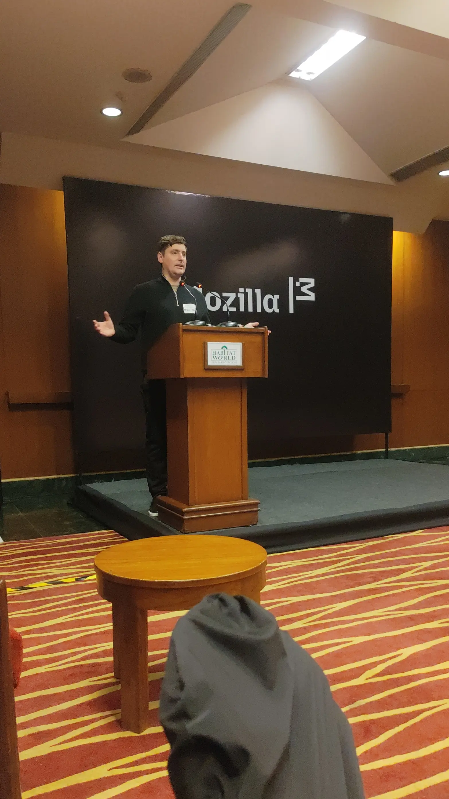 Photogragh of an audience view of John Dickerson giving a talk on a podium on a flat stage with the Mozilla logo banner behind him indoors.