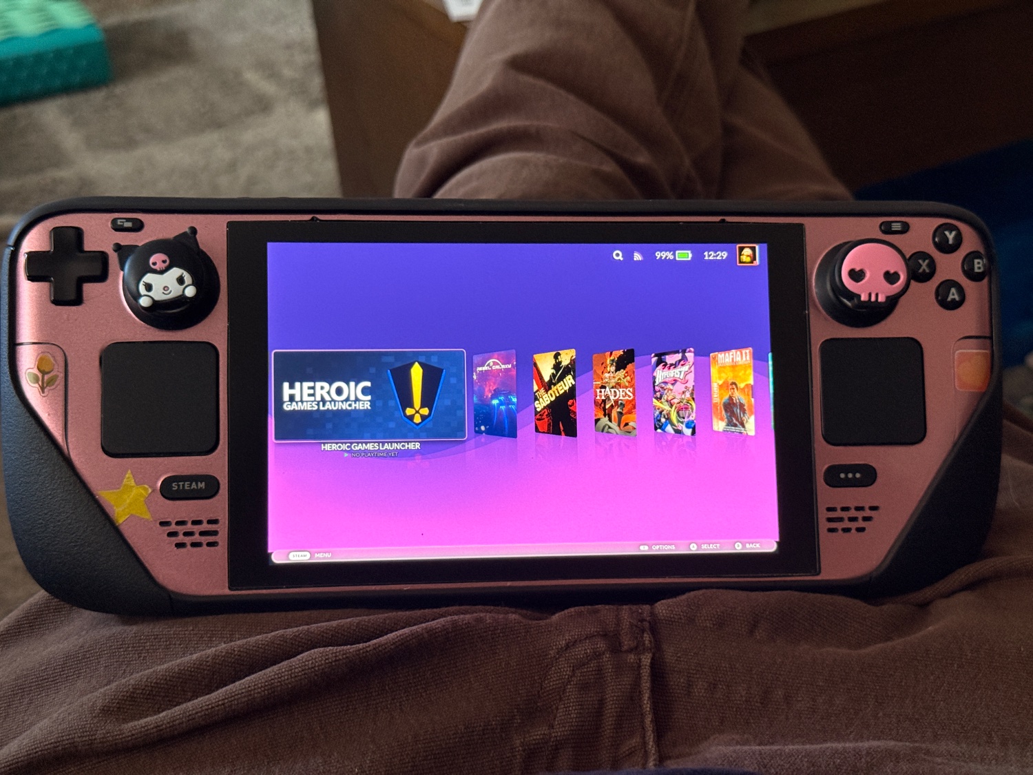 Pink steamdeck with a black protective case around it. There are three stickers on it. A flower, a star, and a heart. There are two silicon thumbstick covers. One is kuromi from hello kitty and the other is a cute pink skull. It is the image of peak gaming. 